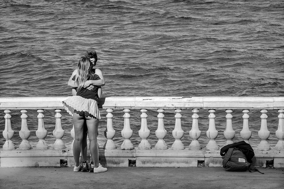A couple embraces beside a balustrade overlooking the sea.